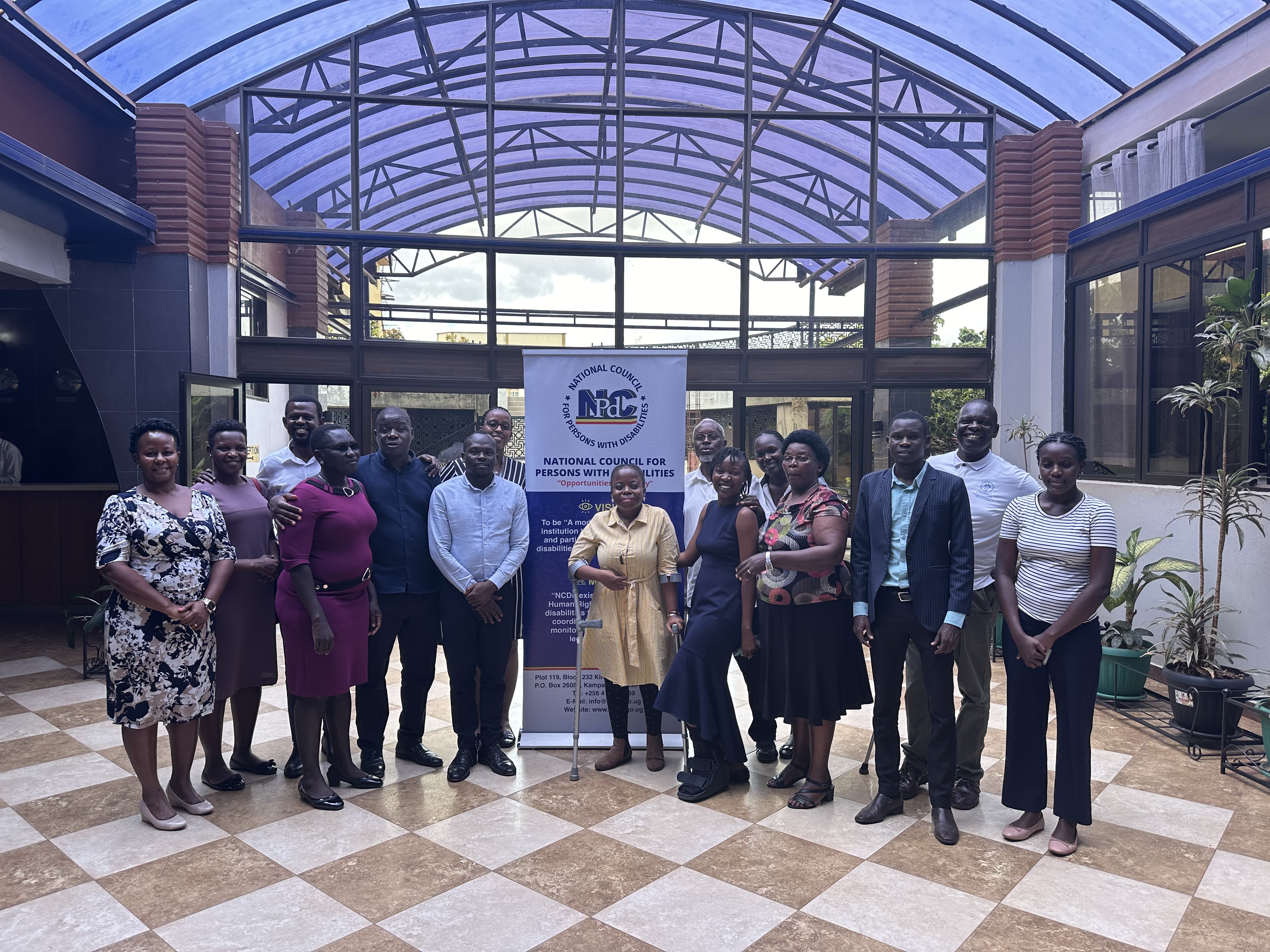 The image features a group of individuals posing together in a well-lit indoor setting, likely during an event related to the National Council for Persons with Disabilities. The backdrop includes a large banner with the council's logo and mission statement.  In the foreground, there are several people standing closely together, smiling for the camera. The group consists of both men and women, dressed in a mix of formal and semi-formal attire. The woman in the center is wearing a light-colored dress and appears to be holding a cane, suggesting she may have a disability.  The setting has a modern design, with a glass roof allowing natural light to filter in. Potted plants are visible in the background, adding a touch of greenery to the environment. The overall atmosphere conveys a sense of community and support among the participants.