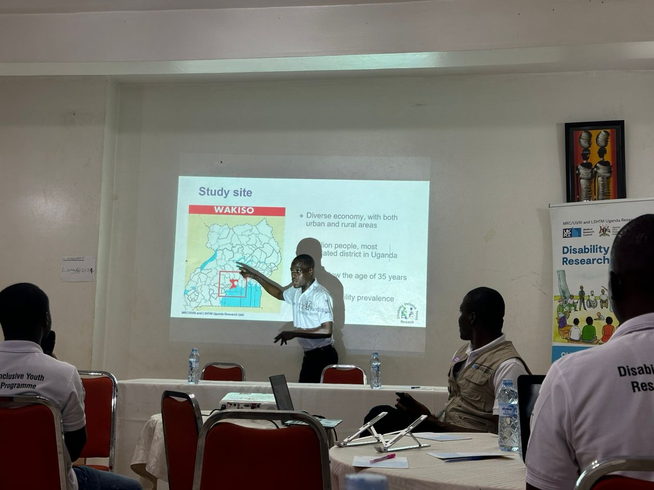 The image depicts a presentation taking place in a conference or workshop setting. A speaker is gesturing towards a slide that highlights the study site, "Wakiso," which is located in Uganda. The slide includes a map showing the area and mentions key points about the region, such as its diverse economy encompassing both urban and rural areas, and demographic details about the population, including a focus on individuals under the age of 35 and disability prevalence.  In the foreground, attendees are seated at tables, some wearing matching shirts that indicate their involvement in a program related to disability research. The atmosphere appears collaborative and educational, with materials and a projector visible, suggesting an interactive session aimed at sharing research findings or methodologies.