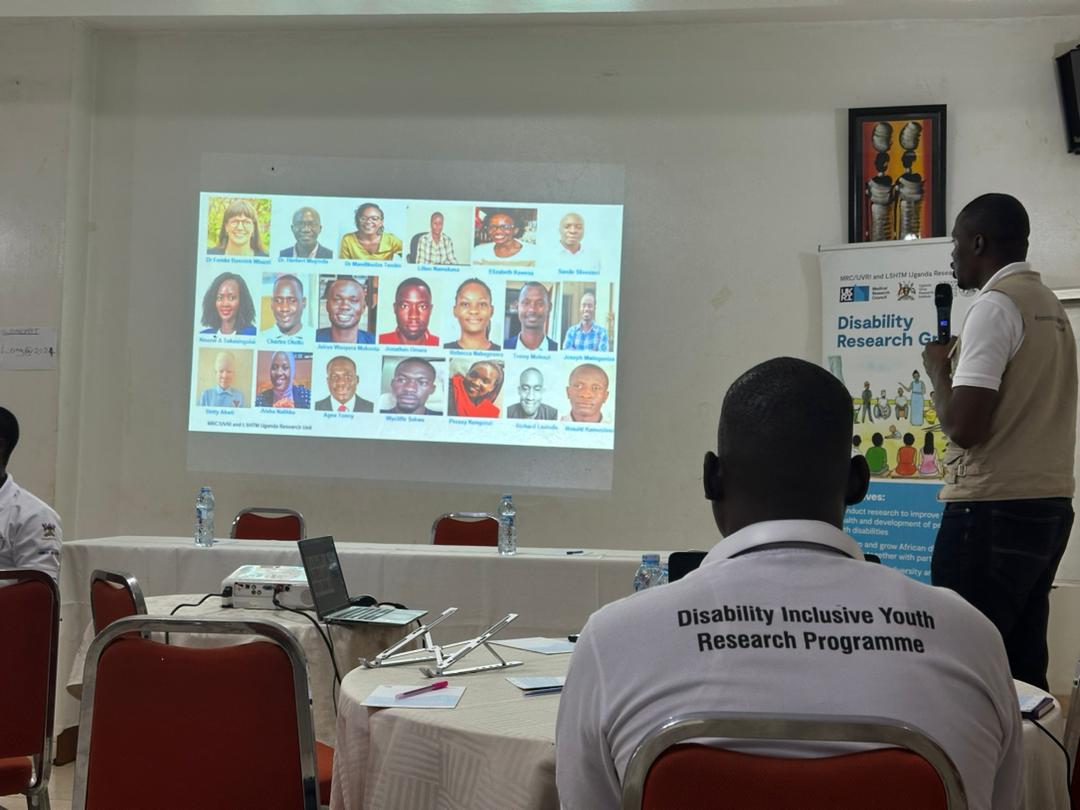 The image depicts a presentation taking place in a conference or workshop setting. A speaker, standing and holding a microphone, is addressing an audience while pointing to a projected slide that features a grid of headshots of various individuals. The slide likely showcases participants or contributors related to a specific project or research initiative.  In the foreground, a person is seated with their back to the camera, wearing a white shirt that reads "Disability Inclusive Youth Research Programme." The setting appears to be organized, with tables set up for attendees, and there are water bottles and a laptop on the table. A banner in the background indicates that the event is focused on disability research, emphasizing the importance of inclusive practices in research and development.