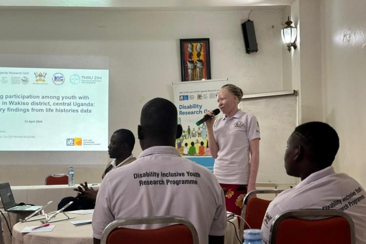 The image depicts a presentation taking place in a conference or workshop setting. A woman is speaking into a microphone, likely presenting findings related to youth participation among individuals with disabilities in Wakiso district, central Uganda. The presentation is supported by a slide displayed on a screen, which includes the title of the research and the date, April 12, 2024.  In the foreground, several attendees are seated at a table, wearing matching shirts that indicate their involvement in a "Di