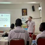 The image depicts a presentation taking place in a conference or workshop setting. A woman is speaking into a microphone, likely presenting findings related to youth participation among individuals with disabilities in Wakiso district, central Uganda. The presentation is supported by a slide displayed on a screen, which includes the title of the research and the date, April 12, 2024.  In the foreground, several attendees are seated at a table, wearing matching shirts that indicate their involvement in a "Di